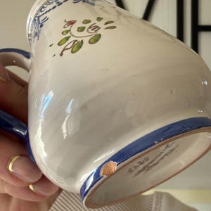 May include: White ceramic pitcher with blue handle and trim. The pitcher features a hand-painted floral design in green, red, and blue. The pitcher has a chip on the bottom rim. A gold ring is visible.