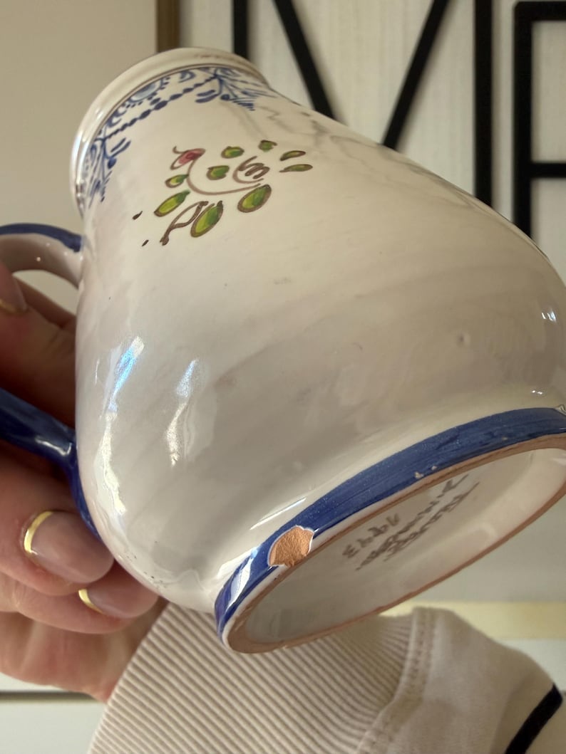 May include: White ceramic pitcher with blue handle and trim. The pitcher features a hand-painted floral design in green, red, and blue. The pitcher has a chip on the bottom rim. A gold ring is visible.