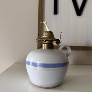 May include: A white ceramic oil lamp with a blue stripe, brass top, and a handle. The lamp has a round body and a wick. The lamp is on a white surface.