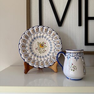 May include: A white ceramic plate with a scalloped edge and floral design, displayed on a wooden stand. A matching mug with a blue handle and floral accents sits beside it. The plate and mug have a blue and yellow color scheme.