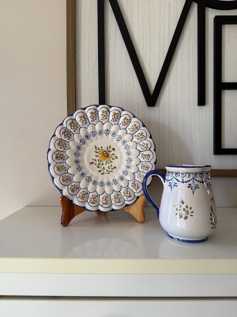 May include: A white ceramic plate with a scalloped edge and floral design, displayed on a wooden stand. A matching mug with a blue handle and floral accents sits beside it. The plate and mug have a blue and yellow color scheme.