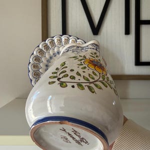 May include: A white ceramic pitcher with a blue rim and floral design. The pitcher features a yellow flower with green leaves and stems. A matching plate with a blue and white pattern is behind the pitcher. The pitcher is held by a person.