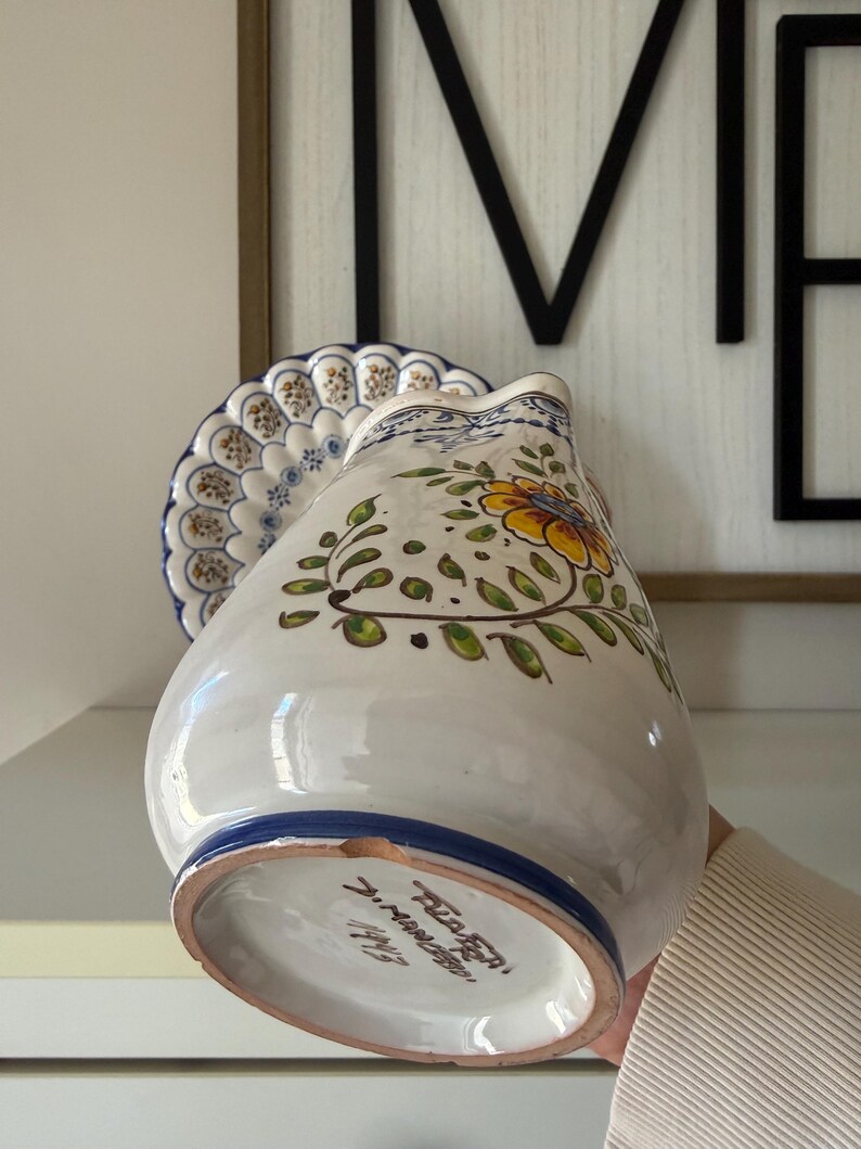 May include: A white ceramic pitcher with a blue rim and floral design. The pitcher features a yellow flower with green leaves and stems. A matching plate with a blue and white pattern is behind the pitcher. The pitcher is held by a person.