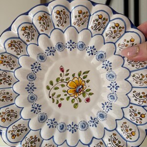 May include: A white ceramic plate with a scalloped edge, adorned with hand-painted floral and leaf motifs in yellow, blue, and green. The plate features a central floral design and a blue border, showcasing a traditional decorative style.