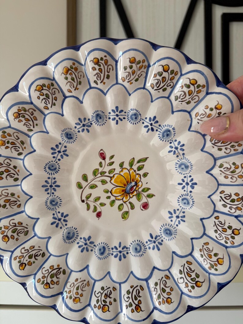 May include: A white ceramic plate with a scalloped edge, adorned with hand-painted floral and leaf motifs in yellow, blue, and green. The plate features a central floral design and a blue border, showcasing a traditional decorative style.