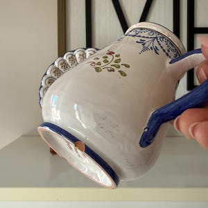 May include: White ceramic pitcher with blue and green floral designs. The pitcher has a blue handle and rim, and a scalloped edge. The pitcher is decorated with a floral pattern and a small brown accent.