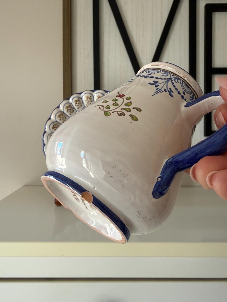 May include: White ceramic pitcher with blue and green floral designs. The pitcher has a blue handle and rim, and a scalloped edge. The pitcher is decorated with a floral pattern and a small brown accent.