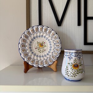 May include: A decorative ceramic plate and vase set. The plate is white with a scalloped edge, blue trim, and floral designs. The vase is white with a similar floral pattern and blue accents. The set is displayed on a white surface.