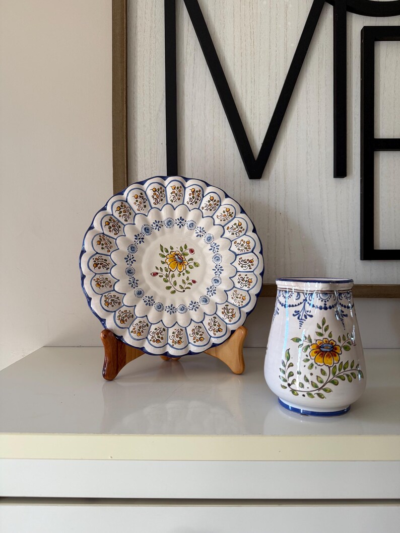 May include: A decorative ceramic plate and vase set. The plate is white with a scalloped edge, blue trim, and floral designs. The vase is white with a similar floral pattern and blue accents. The set is displayed on a white surface.