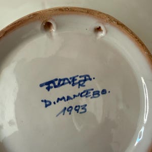 May include: A white ceramic plate with a brown rim and two holes for hanging. The plate has blue handwritten text that reads "TIDER. D.MANCEBO. 1993".