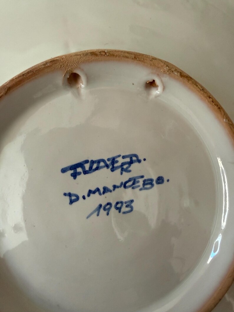 May include: A white ceramic plate with a brown rim and two holes for hanging. The plate has blue handwritten text that reads "TIDER. D.MANCEBO. 1993".