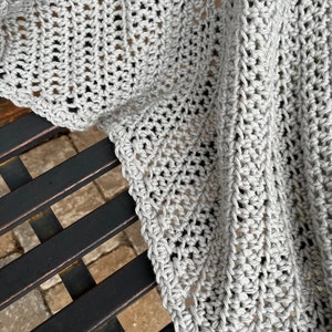 May include: A gray crocheted blanket with a textured pattern. The blanket is draped over a black metal bench.