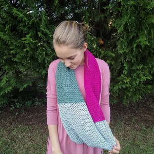 Puede incluir: Una bufanda infinita de color turquesa, gris y rosa hecha de hilo de ganchillo.