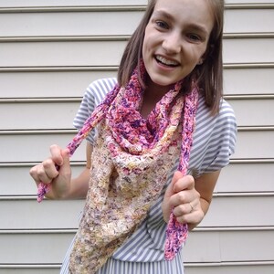 May include: A woman wearing a striped dress and a crocheted shawl in shades of pink, purple, and yellow. The shawl is tied around her neck and the ends are held in her hands.