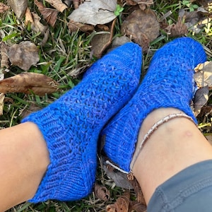 May include: A pair of blue knitted socks with a lacy pattern. The socks are worn on a person's feet and are resting on a bed of green grass and brown leaves.