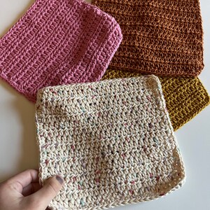May include: Four crocheted dishcloths in different colours: pink, brown, white with multi-coloured flecks, and yellow.