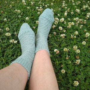 May include: A pair of light blue knitted socks with a textured pattern, resting on a bed of green clover.