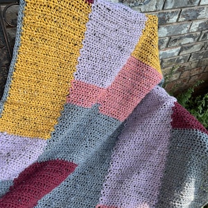 Könnte beinhalten: Eine gehäkelte Decke mit einem geometrischen Muster in Grautönen, Lila, Rosa und Gelb.