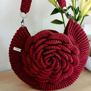 Può includere: Una borsa a mano all'uncinetto bordeaux a forma di rosa. La borsa è dotata di una lunga tracolla e di una chiusura argentata. La borsa fatta a mano ha un'etichetta con la scritta "handmade". La borsa è su una superficie di legno.