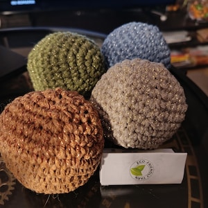 May include: Four crocheted spheres in shades of brown, green, blue, and tan, each with a metallic gold thread woven throughout. The spheres are arranged together, with a small white box that reads "Eco-Friendly Yarn".