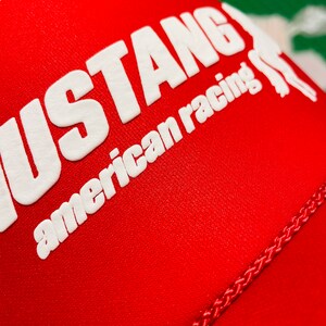 Op de afbeelding: Een felrode honkbalpet met de witte reli&euml;ftekst MUSTANG en american racing, samen met een gedeeltelijke witte afbeelding van een paard. De stof heeft een zichtbare mesh-textuur en een gevlochten rode koordafwerking over het voorpaneel.