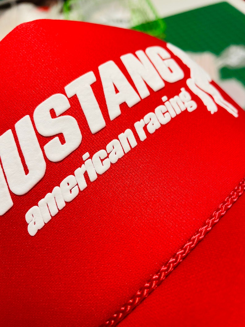 Op de afbeelding: Een felrode honkbalpet met de witte reli&euml;ftekst MUSTANG en american racing, samen met een gedeeltelijke witte afbeelding van een paard. De stof heeft een zichtbare mesh-textuur en een gevlochten rode koordafwerking over het voorpaneel.