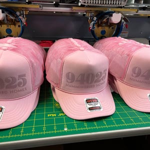 Op de afbeelding: Drie lichtroze truckerpetten met een mesh achterkant en een schuimrubberen voorkant. Elke pet heeft de tekst 94025 en HUMMINGBIRD HOMES op het voorpaneel gedrukt. Een vierkante sticker van het merk OTTO zit op de gebogen klep.