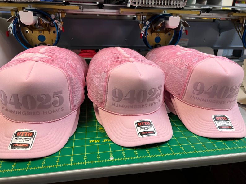 Op de afbeelding: Drie lichtroze truckerpetten met een mesh achterkant en een schuimrubberen voorkant. Elke pet heeft de tekst 94025 en HUMMINGBIRD HOMES op het voorpaneel gedrukt. Een vierkante sticker van het merk OTTO zit op de gebogen klep.