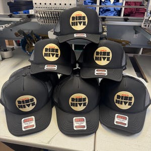 May include: A stack of black trucker hats with a mesh back. Each hat features a circular logo with the words "RISE HIVE" in a gold color. The hats have a tag that says "OTTO TRUCKER".