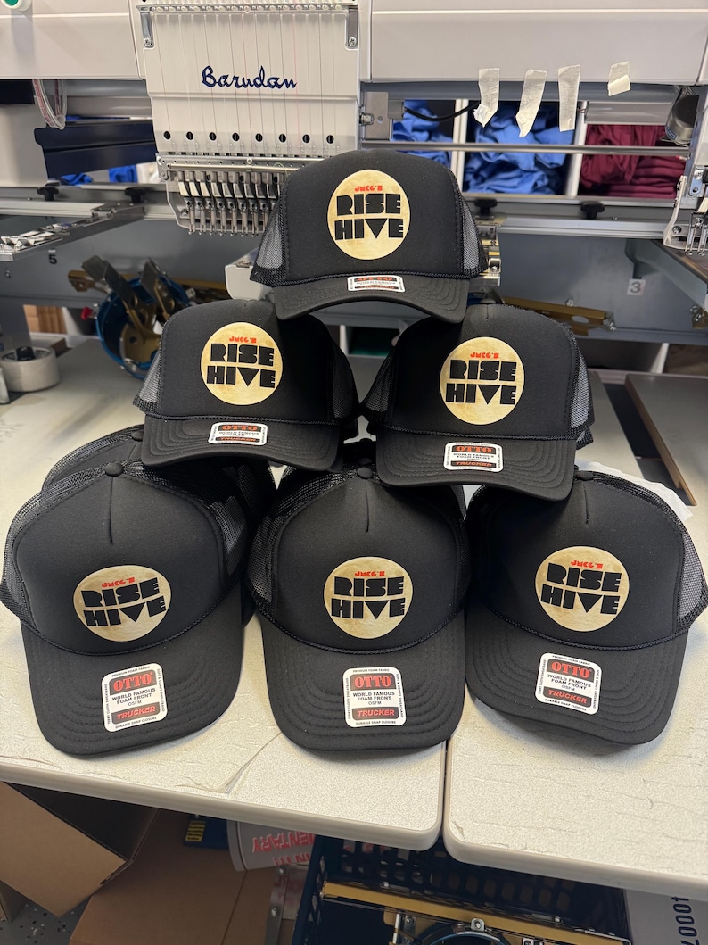 May include: A stack of black trucker hats with a mesh back. Each hat features a circular logo with the words "RISE HIVE" in a gold color. The hats have a tag that says "OTTO TRUCKER".