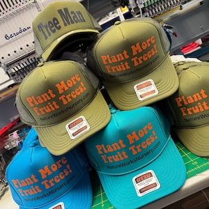 May include: A collection of trucker hats in various colors, including olive green and turquoise. The hats feature the text "Plant More Fruit Trees!" in orange, with "NatureWorksNursery.com" below. One hat reads "Tree Man". Each hat has a mesh back and a front panel.