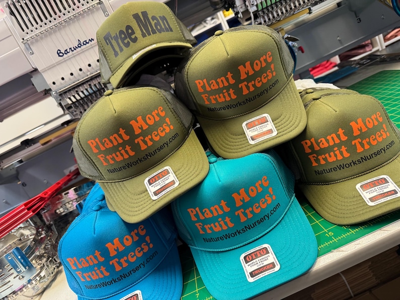 May include: A collection of trucker hats in various colors, including olive green and turquoise. The hats feature the text "Plant More Fruit Trees!" in orange, with "NatureWorksNursery.com" below. One hat reads "Tree Man". Each hat has a mesh back and a front panel.