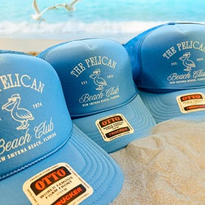 DTF printed OTTO® foam trucker hats in light blue with “The Pelican Beach Club – New Smyrna Beach, Florida” design. Retro coastal vibe, perfect for beach clubs, vacation merch, or family reunions. Sand and sea backdrop.