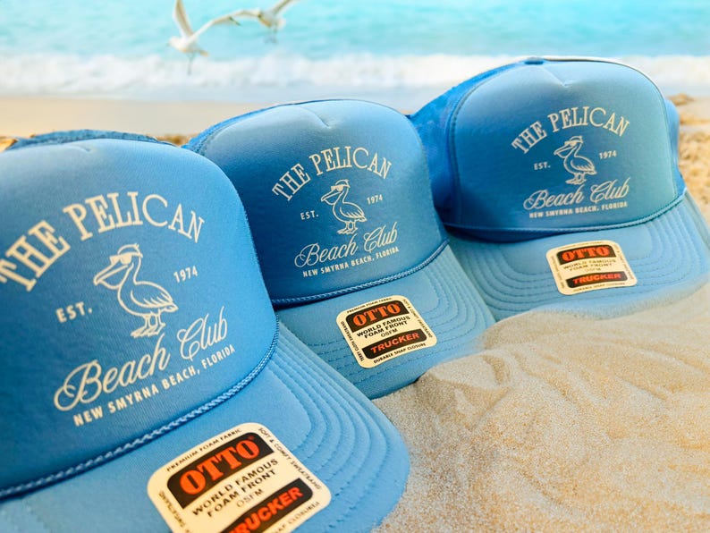 DTF printed OTTO® foam trucker hats in light blue with “The Pelican Beach Club – New Smyrna Beach, Florida” design. Retro coastal vibe, perfect for beach clubs, vacation merch, or family reunions. Sand and sea backdrop.