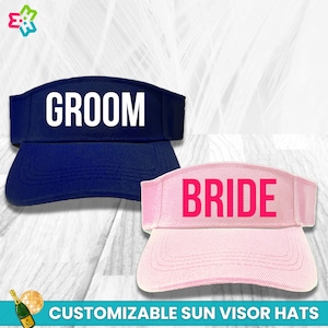 May include: Two customizable sun visor hats, one navy blue with the word "GROOM" in white text and one pink with the word "BRIDE" in pink text.