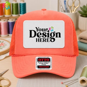 May include: A bright coral trucker hat featuring a white rectangular patch on the front with the black text Your Design Here and a small colorful flower icon. The hat has a foam front, mesh back panels, and a sticker on the brim reading Otto.