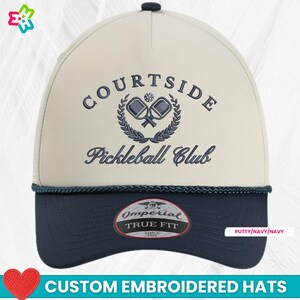 May include: A cream and navy blue Courtside Pickleball Club hat. The hat features embroidered text and a logo of crossed pickleball paddles within a laurel wreath. The bottom of the hat is navy blue, and the text "Custom Embroidered Hats" is at the bottom.