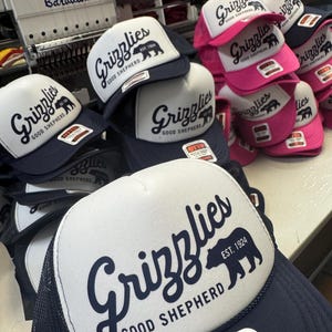May include: A collection of trucker hats with white front panels and navy blue mesh backs. The hats feature the word "Grizzlies" in a script font, a bear silhouette, and the text "Good Shepherd EST. 1924". Some hats have pink brims.