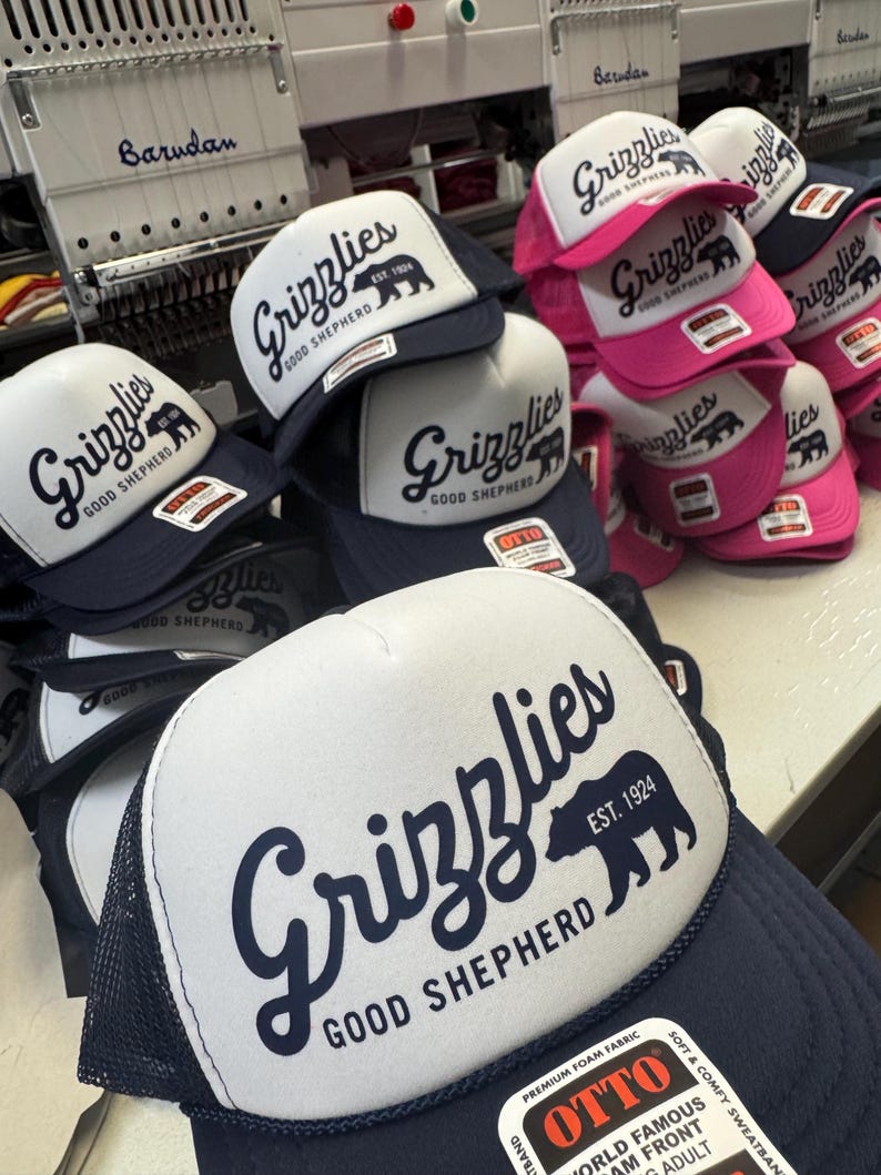 May include: A collection of trucker hats with white front panels and navy blue mesh backs. The hats feature the word "Grizzlies" in a script font, a bear silhouette, and the text "Good Shepherd EST. 1924". Some hats have pink brims.