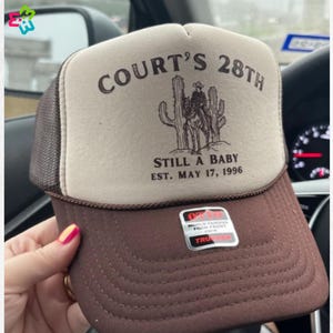 Puede incluir: Gorra de camionero marr&oacute;n y beige con el texto "COURT'S 28TH" sobre un dibujo de un vaquero y cactus. La gorra tambi&eacute;n muestra "STILL A BABY EST. MAY 17, 1996".