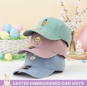 May include: Three pastel-colored dad hats stacked on top of each other. The top hat is mint green with a carrot embroidered on the front. The middle hat is pink with a chick. The bottom hat is light blue with a bunny. The text "EASTER EMBROIDERED DAD HATS" is at the bottom.