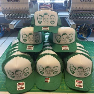 May include: Green and white trucker hats with a screen-printed design of four faces. The hats have a green mesh back and a white front panel. The hats are stacked on a green cutting mat. The brand "OTTO Trucker" is visible on the label.