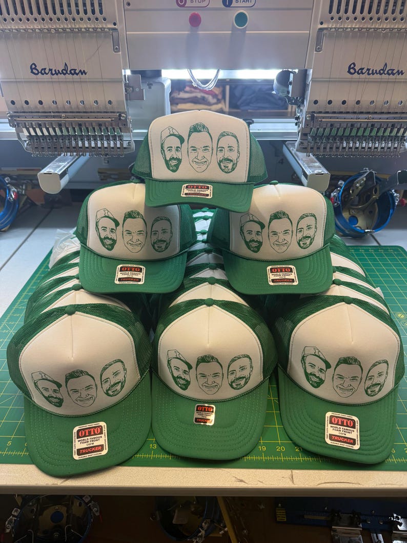 May include: Green and white trucker hats with a screen-printed design of four faces. The hats have a green mesh back and a white front panel. The hats are stacked on a green cutting mat. The brand "OTTO Trucker" is visible on the label.