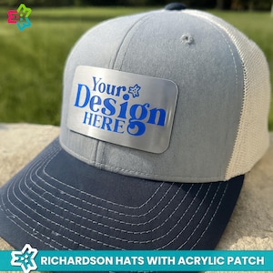 May include: A gray and navy blue trucker hat with a white mesh back. The hat features a silver acrylic patch with the blue text "Your Design HERE". The hat is a Richardson hat.