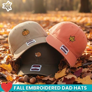May include: Three embroidered dad hats in tan, orange, and olive green, each with a fall-themed design. The tan hat features a pumpkin, the orange hat a maple leaf, and the olive green hat a sleeping fox. The hats are displayed on a bed of autumn leaves.