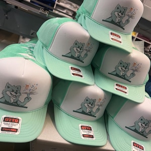 May include: A stack of mint green and white trucker hats. Each hat features a cartoon fox holding a flower and the text "OTTO WORLD FAMOUS FOAM FRONT TRUCKER". The hats have a mesh back and a curved brim.