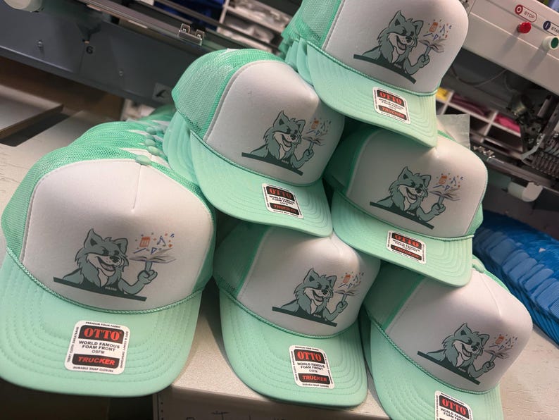 May include: A stack of mint green and white trucker hats. Each hat features a cartoon fox holding a flower and the text "OTTO WORLD FAMOUS FOAM FRONT TRUCKER". The hats have a mesh back and a curved brim.