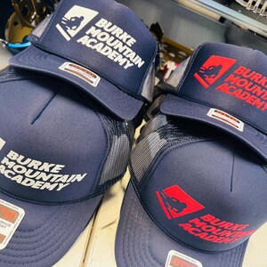 Op de afbeelding: Twee marineblauwe truckerpetten met mesh achterkant en schuimrubberen voorkant. De ene pet heeft witte tekst en een logo met Burke Mountain Academy, de andere rode tekst en een logo met Burke Mountain Academy. Beide hebben Otto-stickers.