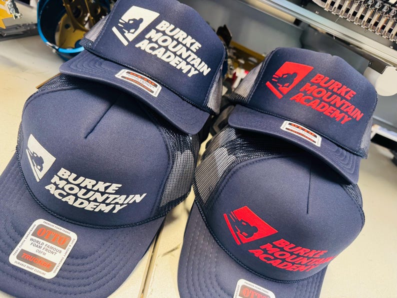 Op de afbeelding: Twee marineblauwe truckerpetten met mesh achterkant en schuimrubberen voorkant. De ene pet heeft witte tekst en een logo met Burke Mountain Academy, de andere rode tekst en een logo met Burke Mountain Academy. Beide hebben Otto-stickers.