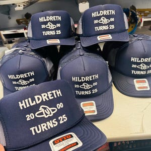 Op de afbeelding: Een collectie marineblauwe truckerpetten met een witte mesh achterkant. Elke pet heeft witte bedrukte tekst met HILDRETH 2000 TURNS 25 en een centrale afbeelding. De petten hebben een gestructureerde schuimrubberen voorkant en een rechthoekige sticker.